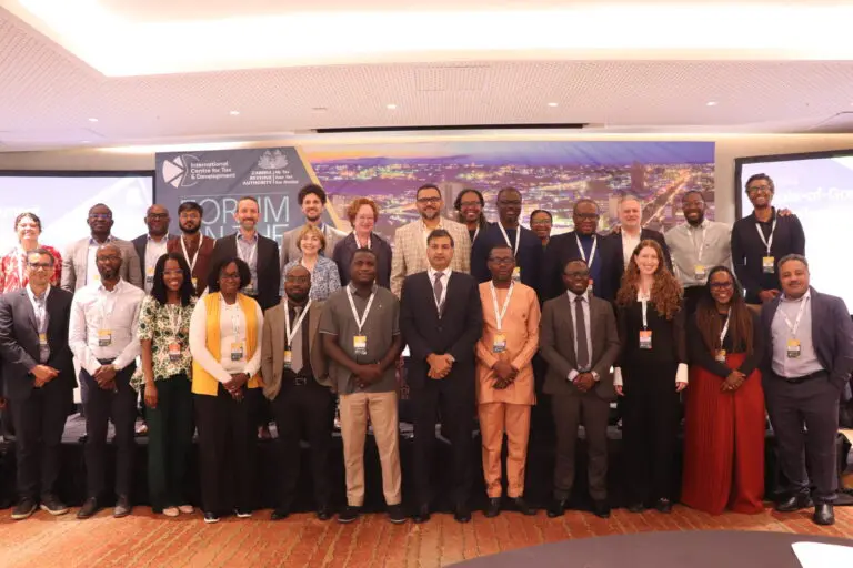 Group photo of diverse professionals standing together at a conference event with badges and a presentation screen in the background.
