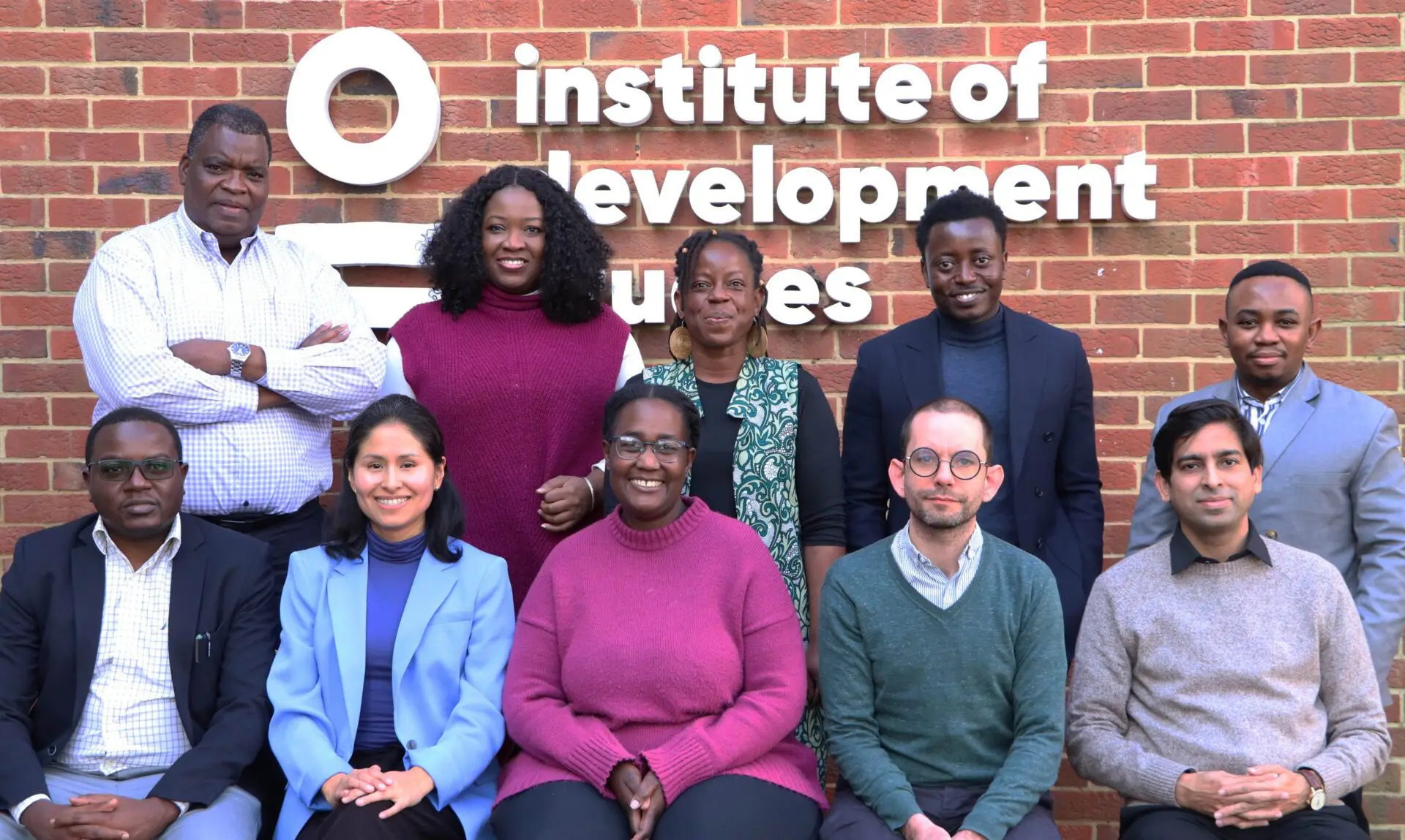 A group of 10 diverse individuals posing for a photo in two levels, five sitting down at the front with the other five standing in the back, in front of the building with a sign that reads "Institute of Development Studies".