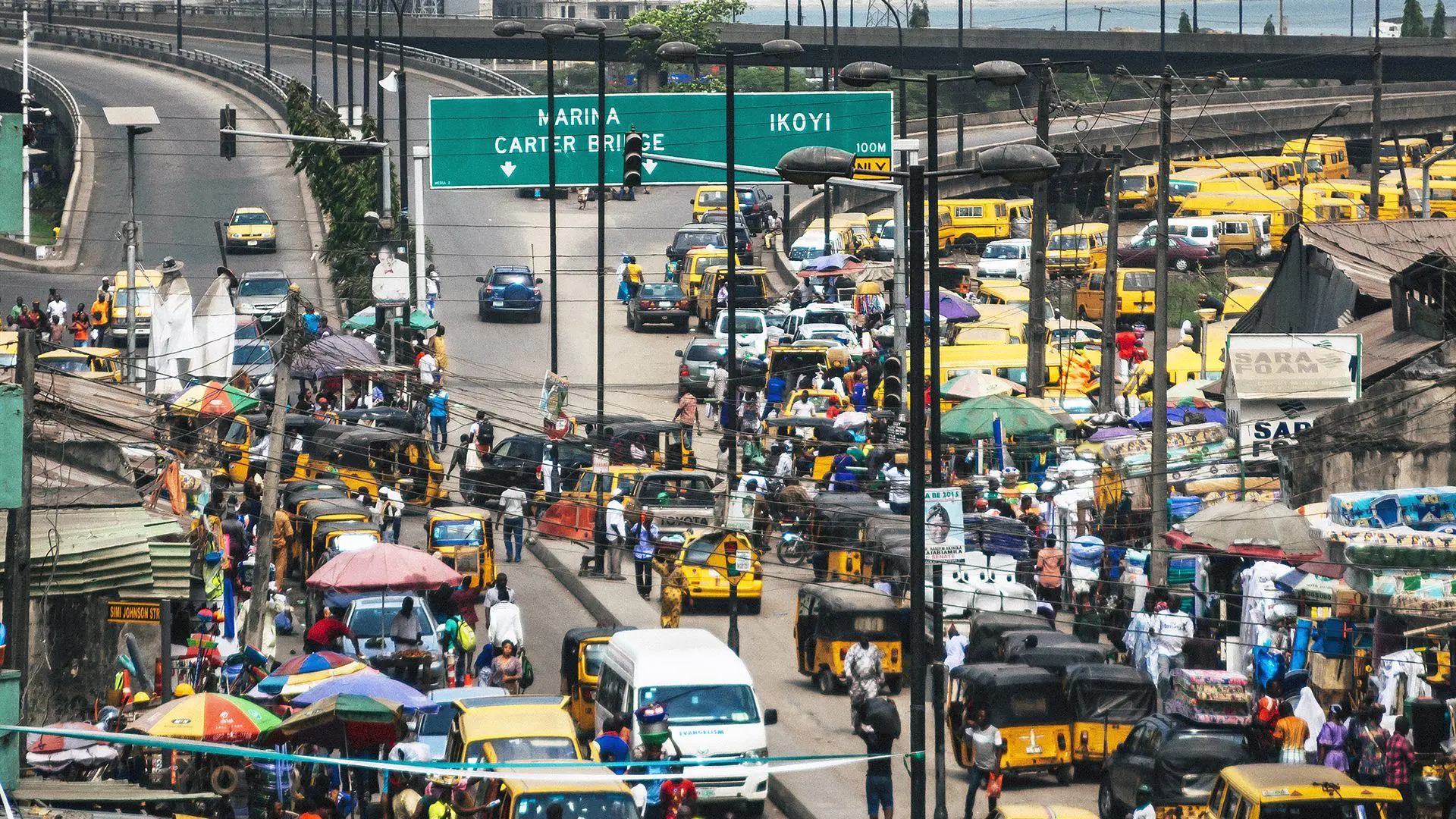 Rue animée de Lagos, avec des minibus jaunes, des étals de marché et un panneau indiquant Marina, Carter Bridge et Ikoyi