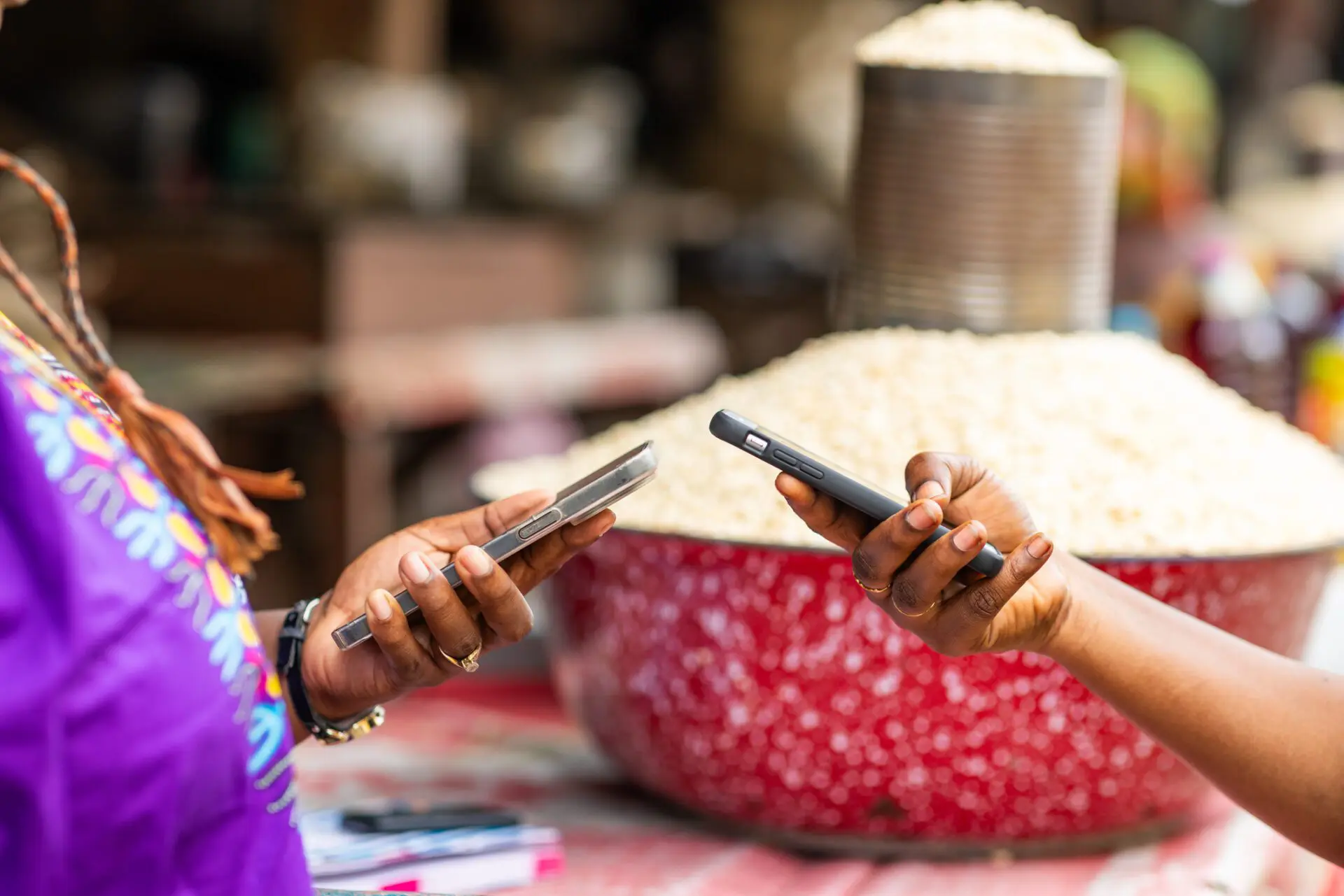 Two women holding smart phones. One is making a digital mobile money payment for fresh grains at vibrant African local outdoor market.