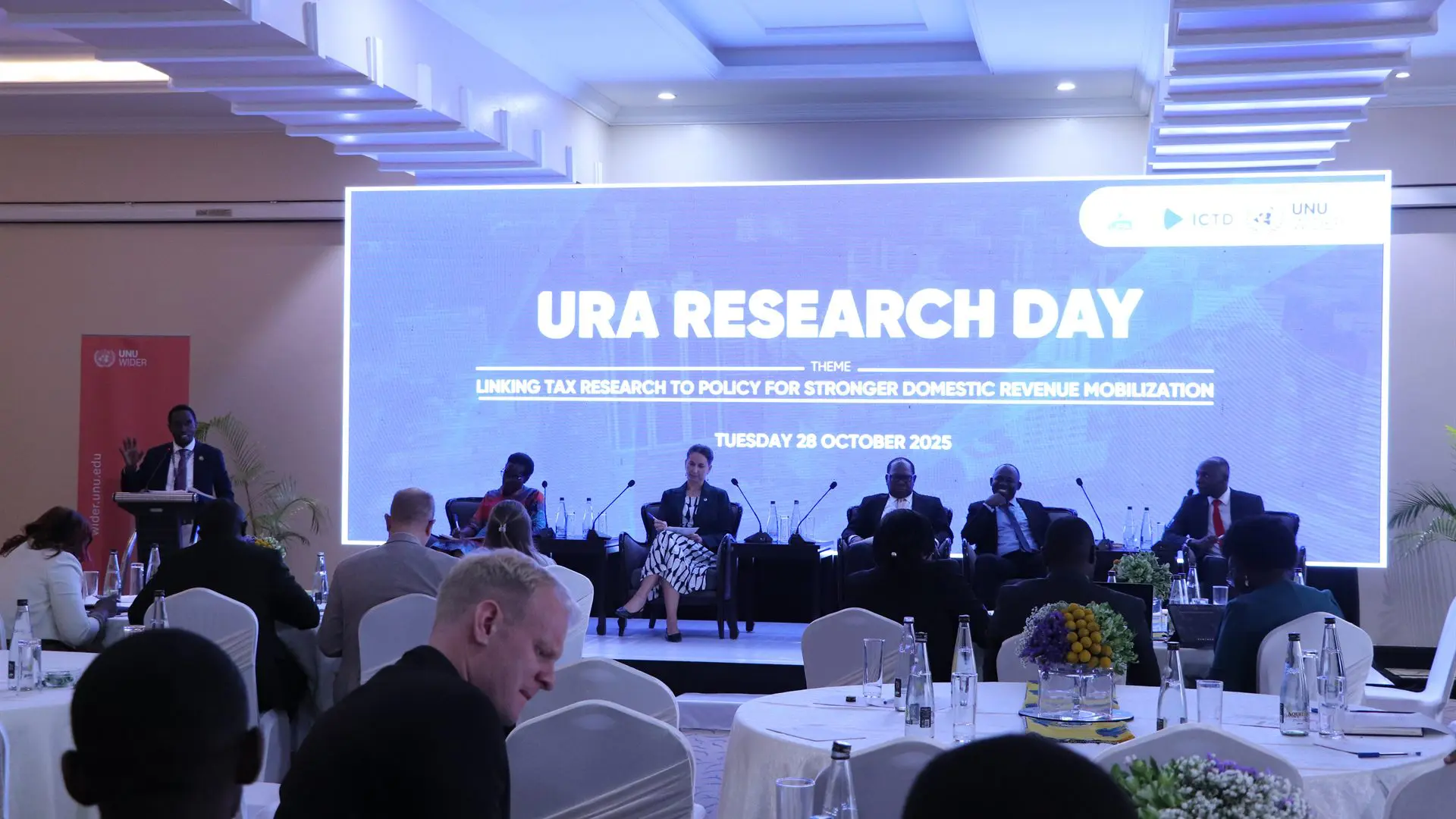 Table ronde sur scène lors de la URA Research Day, avec un orateur au pupitre et des invités assis à des tables rondes