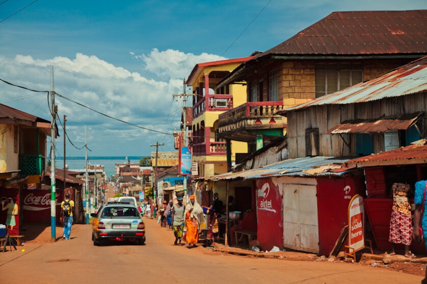 View of a street in Freetown Sierra Leone with buildings on either sides and people walking as a car passes by.