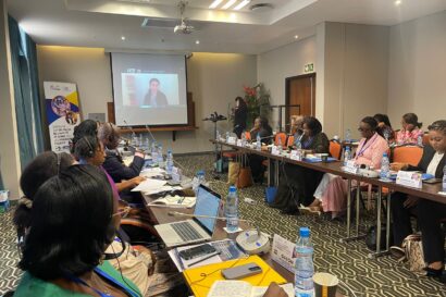 Wide picture showing partially a conference room with people sitting around a U shaped table looking at a projected presentation with a speaker